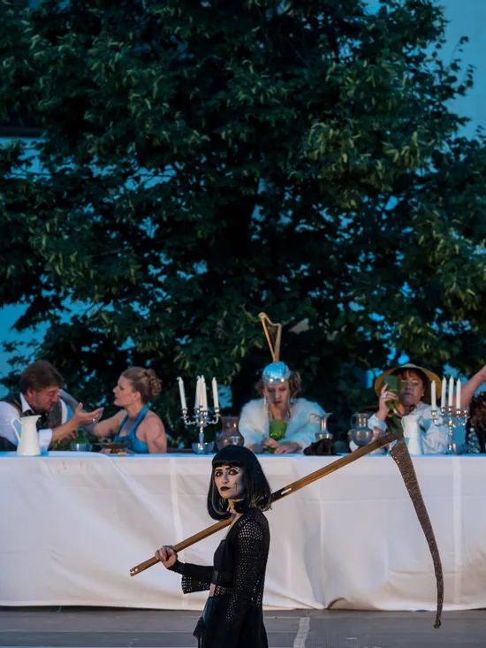 Theatrical outdoor performance with costumed actors at a long dinner table, woman holding scythe in foreground