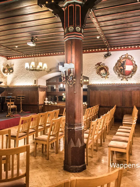 Konzertsaal mit Holzstühlen, Flügel, Bühnenbeleuchtung und mittelalterlichen Wappen