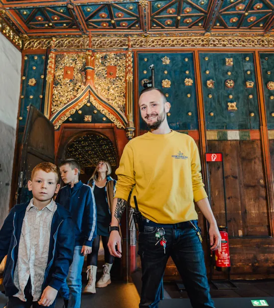 Guida turistica accompagna bambini in una sala storica decorata con pareti e soffitto in legno intarsiato