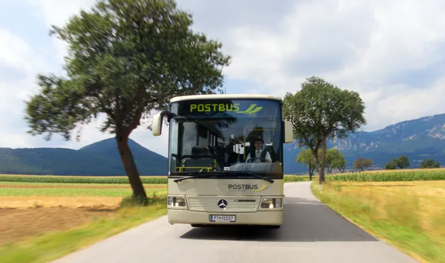 Postbus driving on rural road with trees and mountains in the background