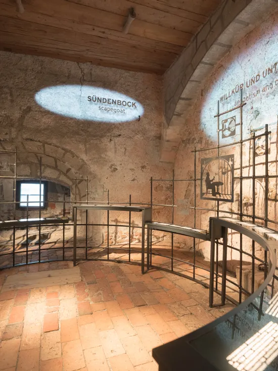 Historische Museumsausstellung über Sündenböcke und Unterdrückung in einem Raum mit Steinwänden und Metallrahmen
