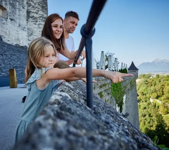 Familie mit Kindern zeigt von einer Burgterrasse ins grüne Tal mit Blick auf entfernte Berge
