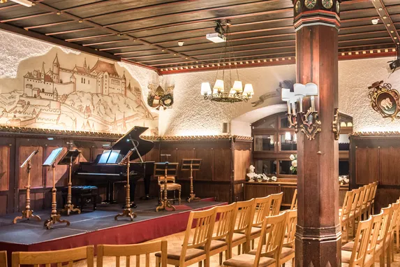 Konzertsaal mit Holzstühlen, Flügel, Bühnenbeleuchtung und mittelalterlichen Wappen