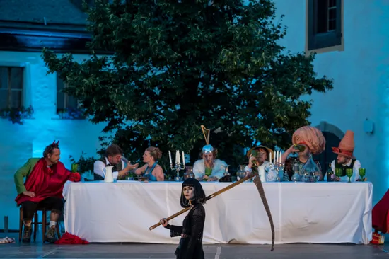 Theatrical outdoor performance with costumed actors at a long dinner table, woman holding scythe in foreground