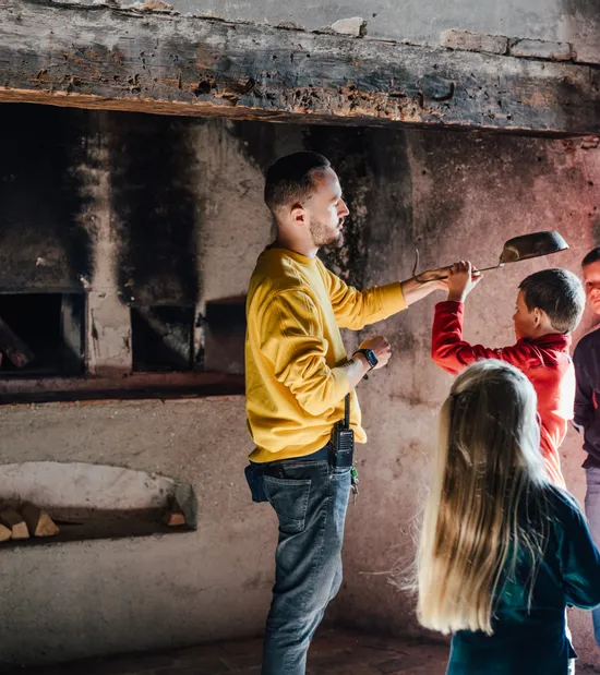 Mann zeigt Kindern den Umgang mit einem Pizzaschieber vor einem holzbefeuerten Backofen